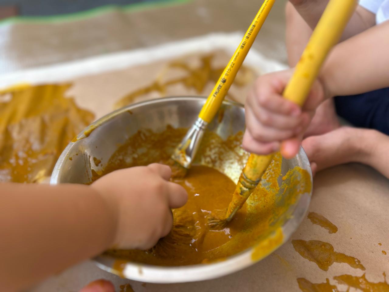 Mãos pequenas de crianças explorando tinta amarela com pincéis durante atividade de expressão artística no Pequeno Urso