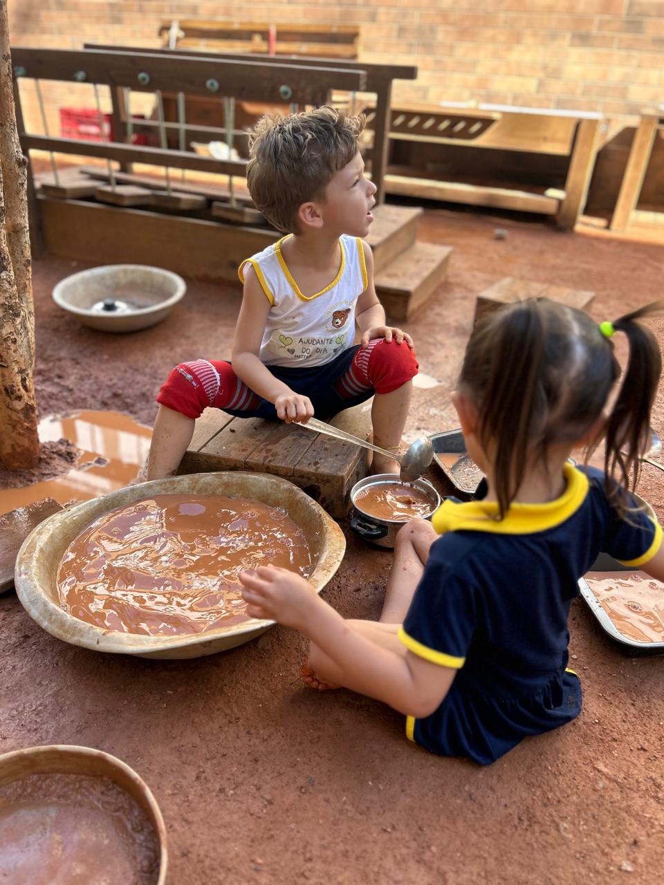 Dois alunos do Pequeno Urso brincando no parque sensorial, explorando água e terra com colheres no pátio da escola