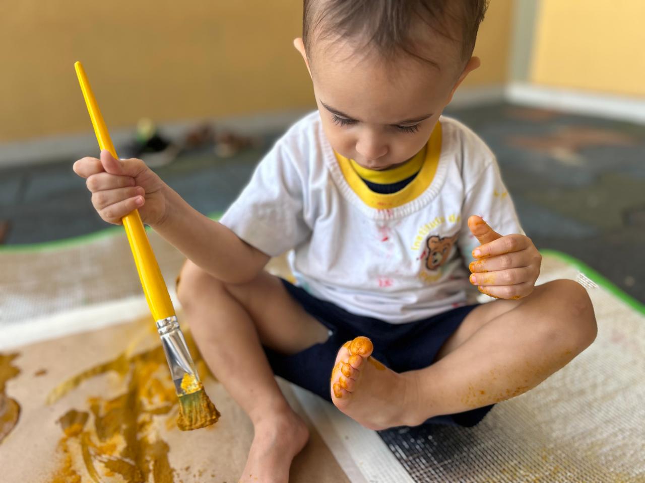 Bebê do Pequeno Urso sentado com pernas cruzadas, concentrado descobrindo tinta amarela em seus pés durante atividade sensorial